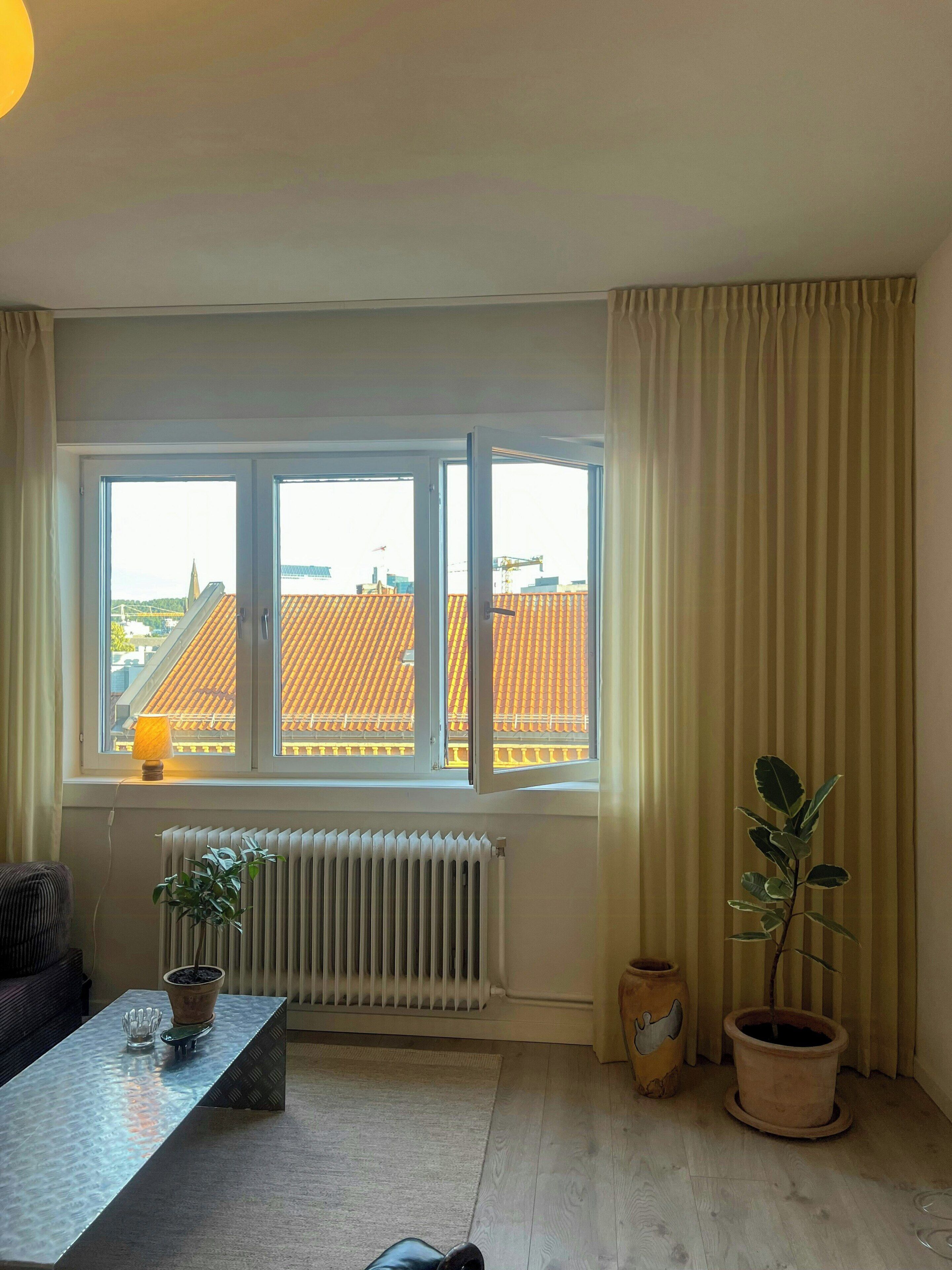 A room with a large window, a radiator, a table with a potted plant, and a potted plant on the floor.