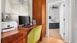 Een houten bureau met een computer, printer en ingelijste foto’s. Een bed en een plafondventilator zijn in de aangrenzende kamer zichtbaar.