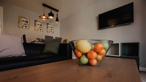 A living room with a wooden coffee table, a glass bowl with fruits, a black sofa, and artwork on the wall. - Coyle House sleeps 7 free parking (Northamptonshire)