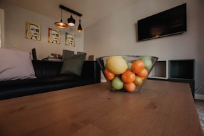 Sala de estar com mesa de centro de madeira, tigela de vidro com frutas, sofá preto e obras de arte na parede.