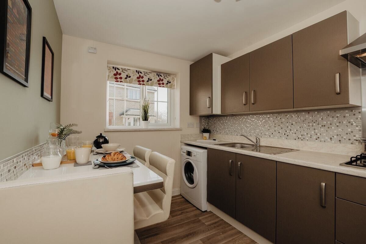 A compact kitchen with a washing machine, a dining area with a table and chairs, and a window with a floral curtain.