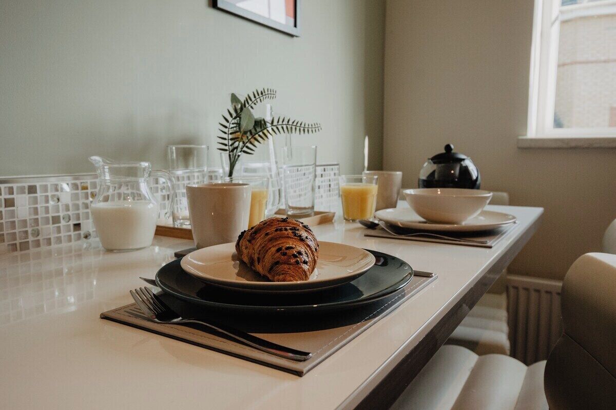 A breakfast setting with a croissant on a plate, a pitcher, a glass, and a kettle on a table.