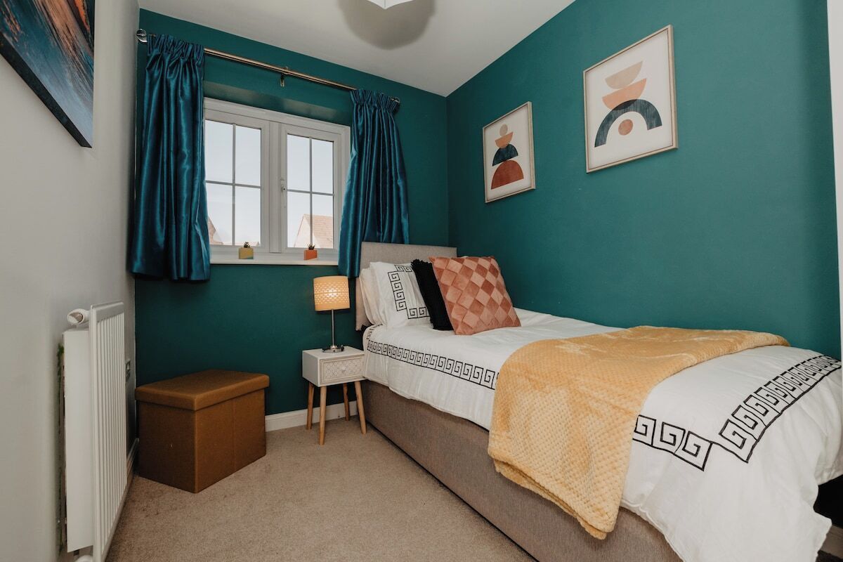 A bedroom with teal walls, a bed with patterned bedding, a bedside table with a lamp, and a window with curtains.