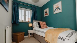 A bedroom with teal walls, a bed with patterned bedding, a bedside table with a lamp, and a window with curtains. - Coyle House sleeps 7 free parking (Northamptonshire)