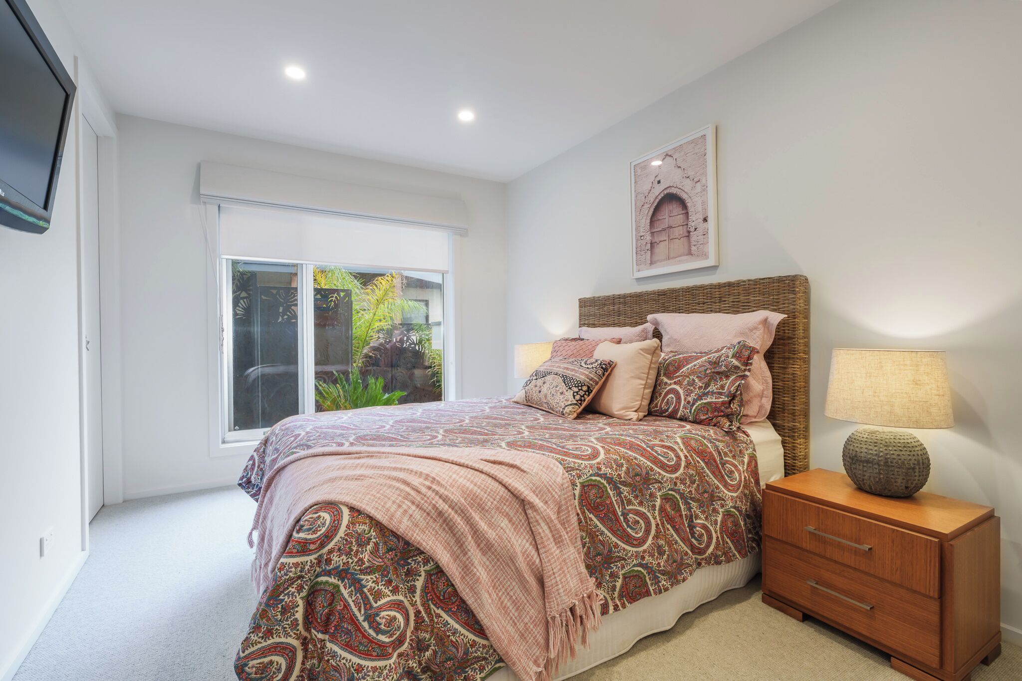 A bedroom with a bed, a nightstand, a lamp, a window, and a wall-mounted picture.