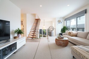 A modern living room with a staircase, a flat-screen TV, a sofa, and a coffee table. - Coastal Haven on Baker (Ocean Grove)