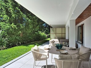 Un espace extérieur aménagé pour prendre les repas, avec une table et des chaises, entouré d’un jardin et offrant une vue sur les arbres.
