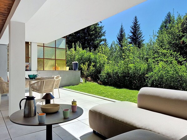 Un espace extérieur moderne, aménagé avec un canapé et une table, une cloison en verre et une vue sur un jardin bien entretenu.