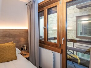 A hotel room with a wooden headboard, a bed with a white comforter, a nightstand with a lamp, a window with wooden frames, and a view of an outdoor area with a table and chairs. - Beautiful apartment for 4 guests with WIFI, TV, balcony, pets allowed and panoramic view (Saint Moritz)