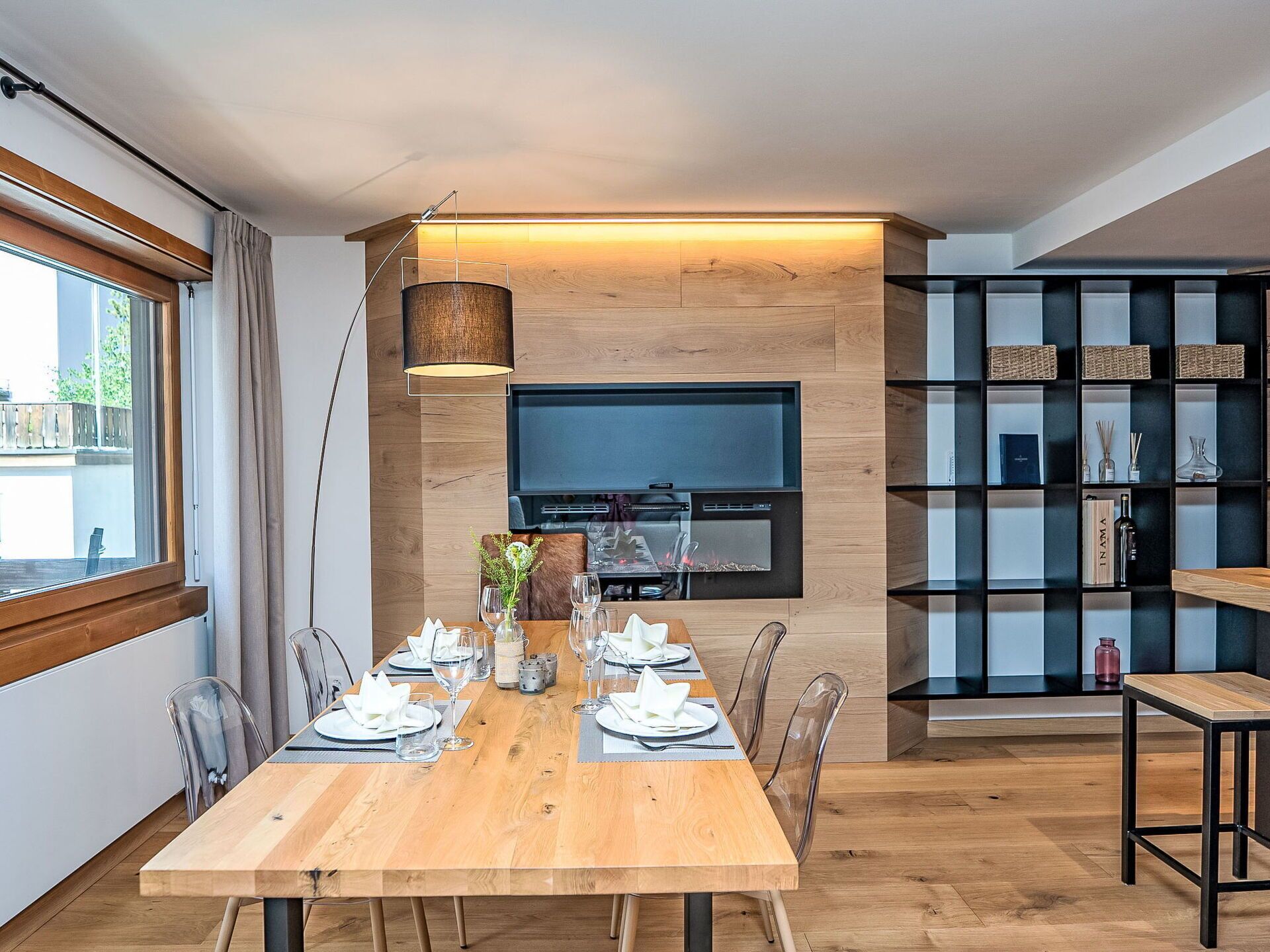 Un comedor moderno con una mesa de madera dispuesta para dos, un televisor montado en una pared de madera y una estantería negra con diversos objetos.