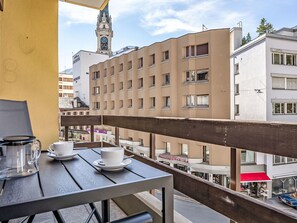 A balcony with a table set for two, overlooking a cityscape with buildings and a clock tower. - Beautiful apartment for 4 guests with WIFI, TV, balcony, pets allowed and panoramic view (Saint Moritz)