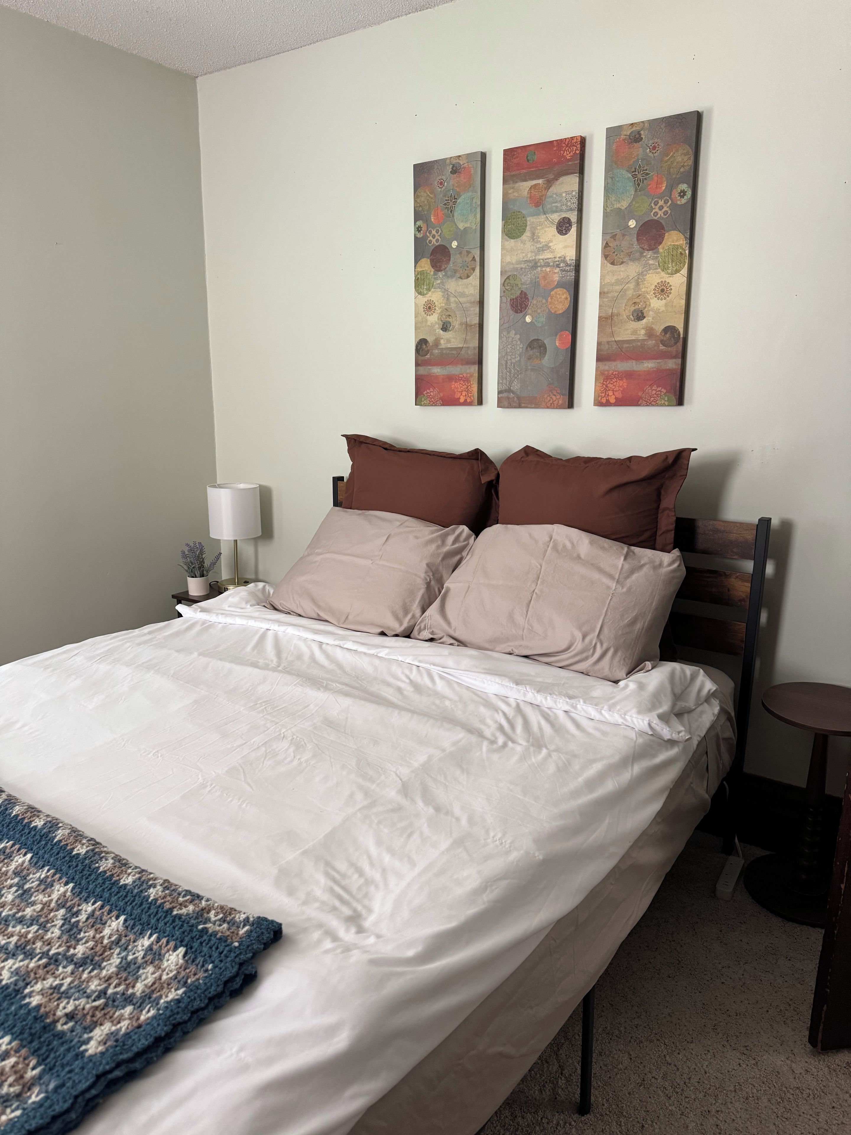 A neatly made bed with a white duvet and two brown pillows, a small bedside table with a lamp, and a wall with three abstract paintings.
