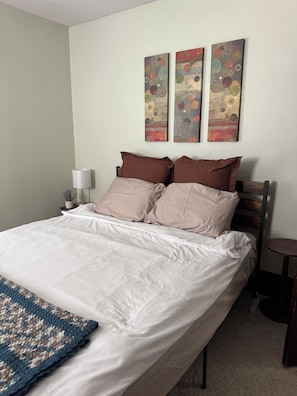 A neatly made bed with a white duvet and two brown pillows, a small bedside table with a lamp, and a wall with three abstract paintings.