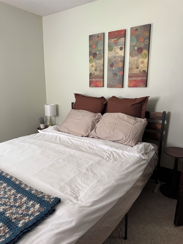 A neatly made bed with a white duvet and two brown pillows, a small bedside table with a lamp, and a wall with three abstract paintings.