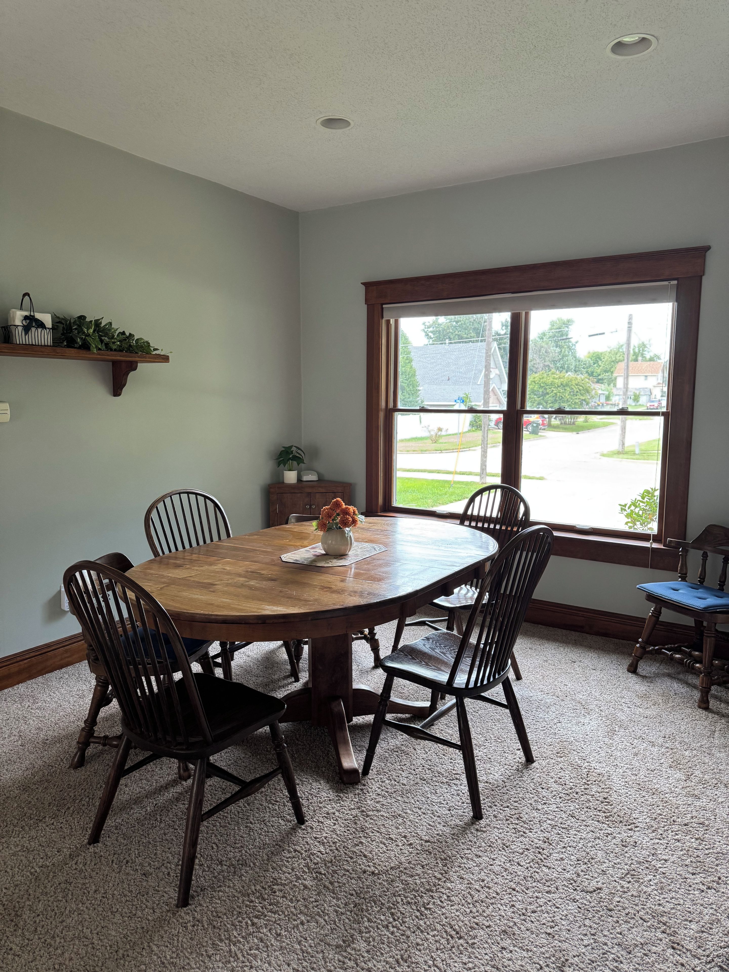 A dining room with a large wooden table, six chairs, a vase with flowers, a window with a view of a street, and a shelf with plants.