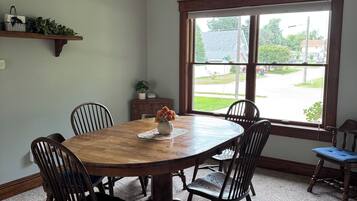 A dining room with a large wooden table, six chairs, a vase with flowers, a window with a view of a street, and a shelf with plants.