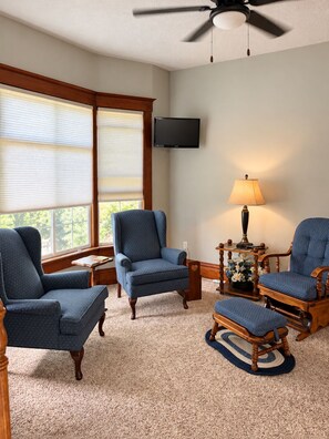 A living room with a ceiling fan, a television mounted on the wall, and a large window with blinds.