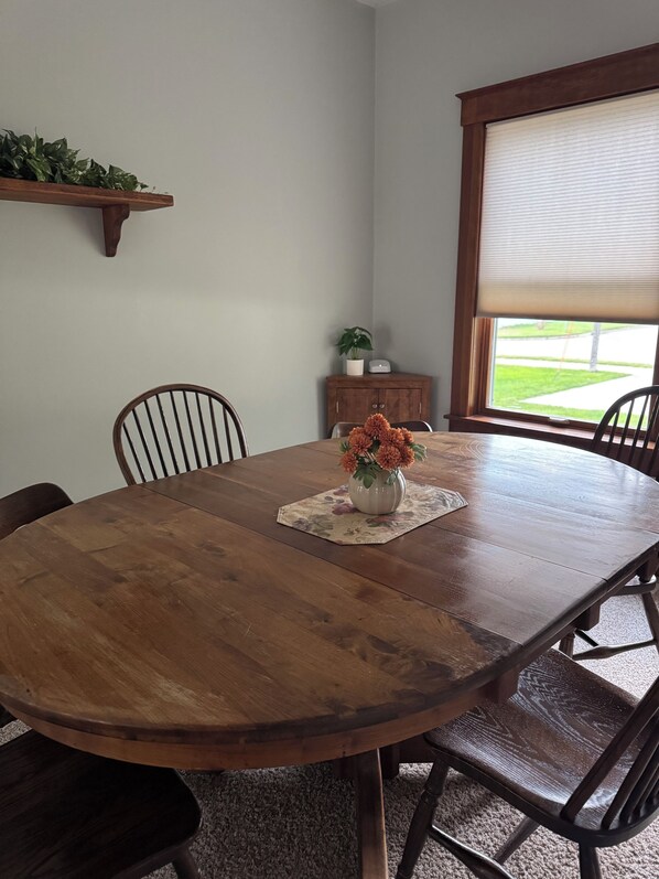 A wooden dining table with chairs, a vase with flowers, a small cabinet, and a window with blinds.