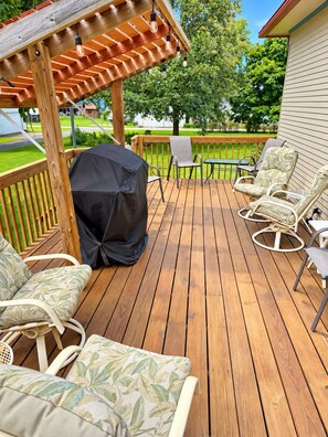 A wooden deck with outdoor furniture, including a covered grill, lounge chairs, and a pergola with hanging lights.