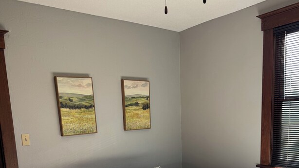 Une chambre à coucher avec un lit, deux tableaux accrochés au mur, un ventilateur de plafond et une fenêtre avec des stores.