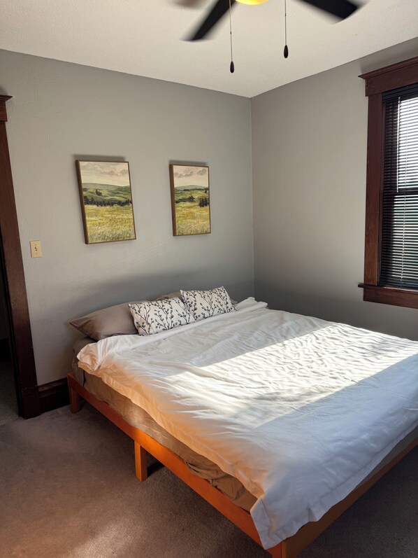 A bedroom with a bed, two wall-mounted paintings, a ceiling fan, and a window with blinds.