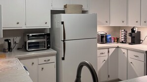 Une cuisine avec des meubles blancs, un four à micro-ondes encastré, un réfrigérateur et une machine à café posée sur le plan de travail.