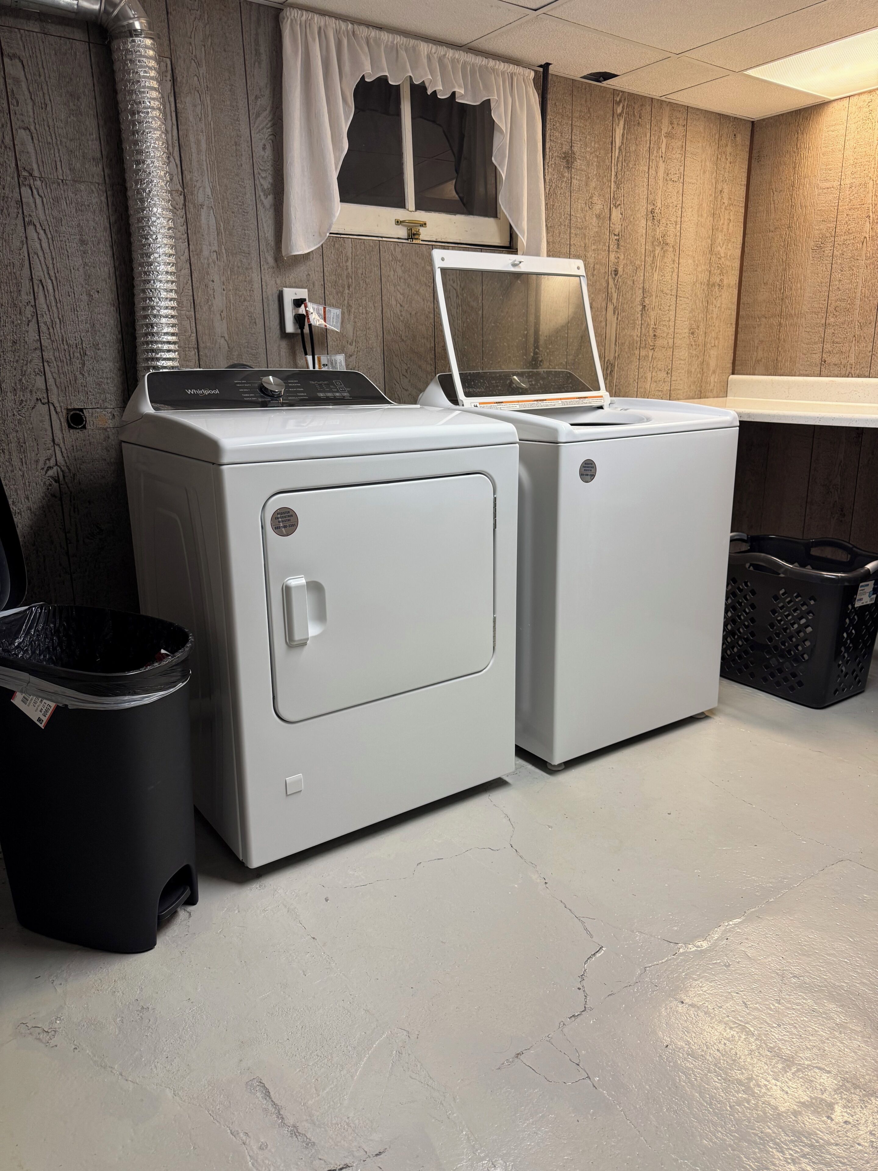 A laundry room with two front-loading washers, a trash bin, and a window with curtains.