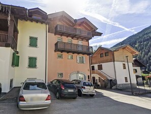 Una fila di edifici a più piani con balconi, auto parcheggiate e montagne sullo sfondo.