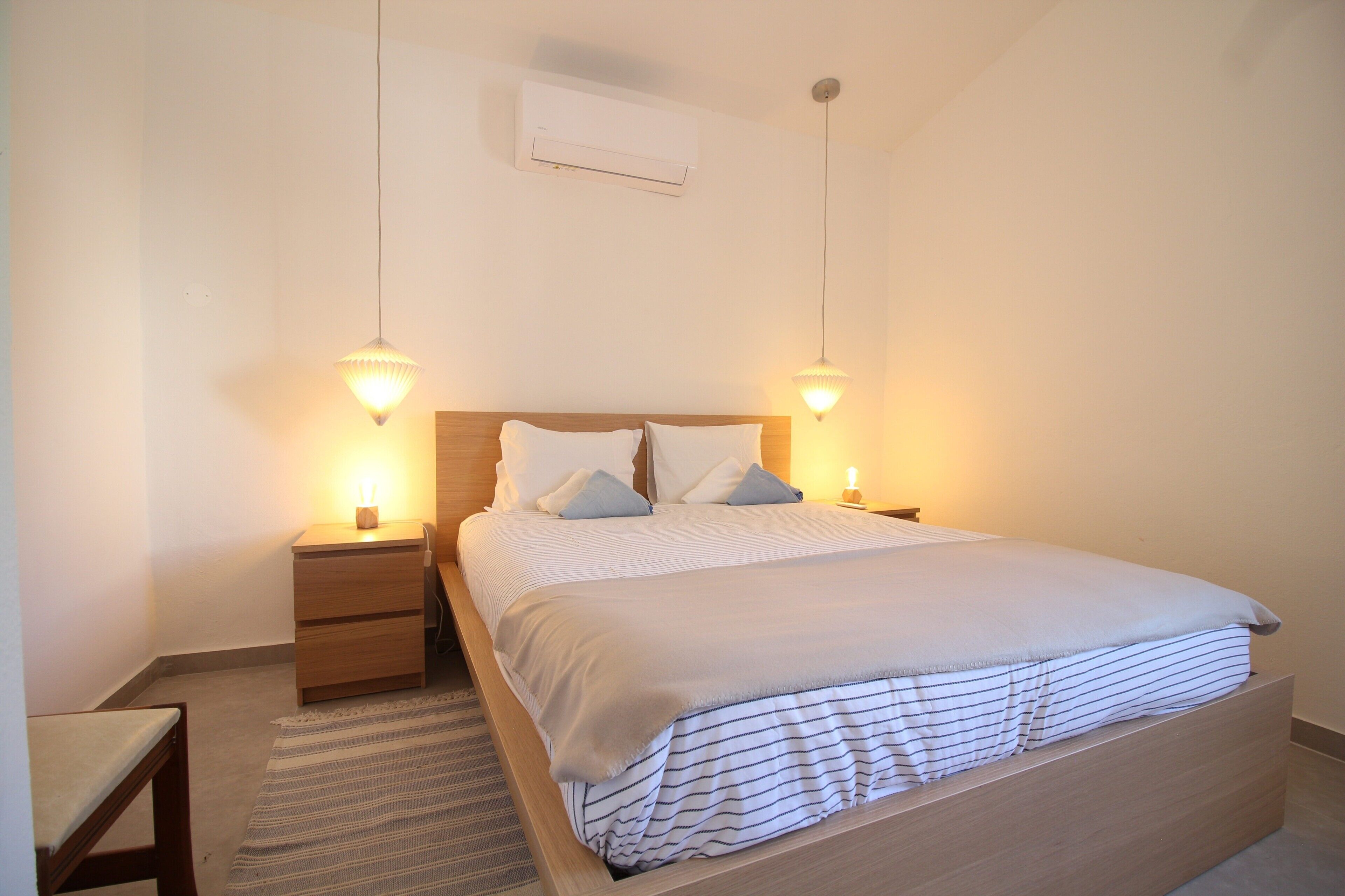 Une chambre à coucher comprenant un lit, deux tables de chevet, une commode, un climatiseur fixé au mur et deux lampes suspendues.