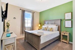 Une chambre à coucher comprenant un lit, des tables de chevet, une lampe, une fenêtre avec des rideaux et un mur orné de tableaux.
