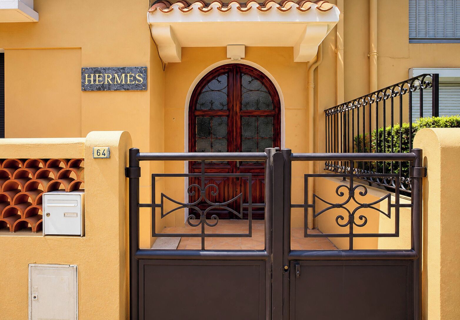 A gated entrance with a sign reading 'HERMES' above a door, a mailbox, and a number '64' on the wall.