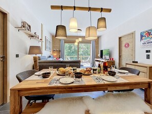 A dining area with a wooden table set for a meal, a sofa, and a television in the background. - Santal - Les Gets - Selena Residence (Les Gets)