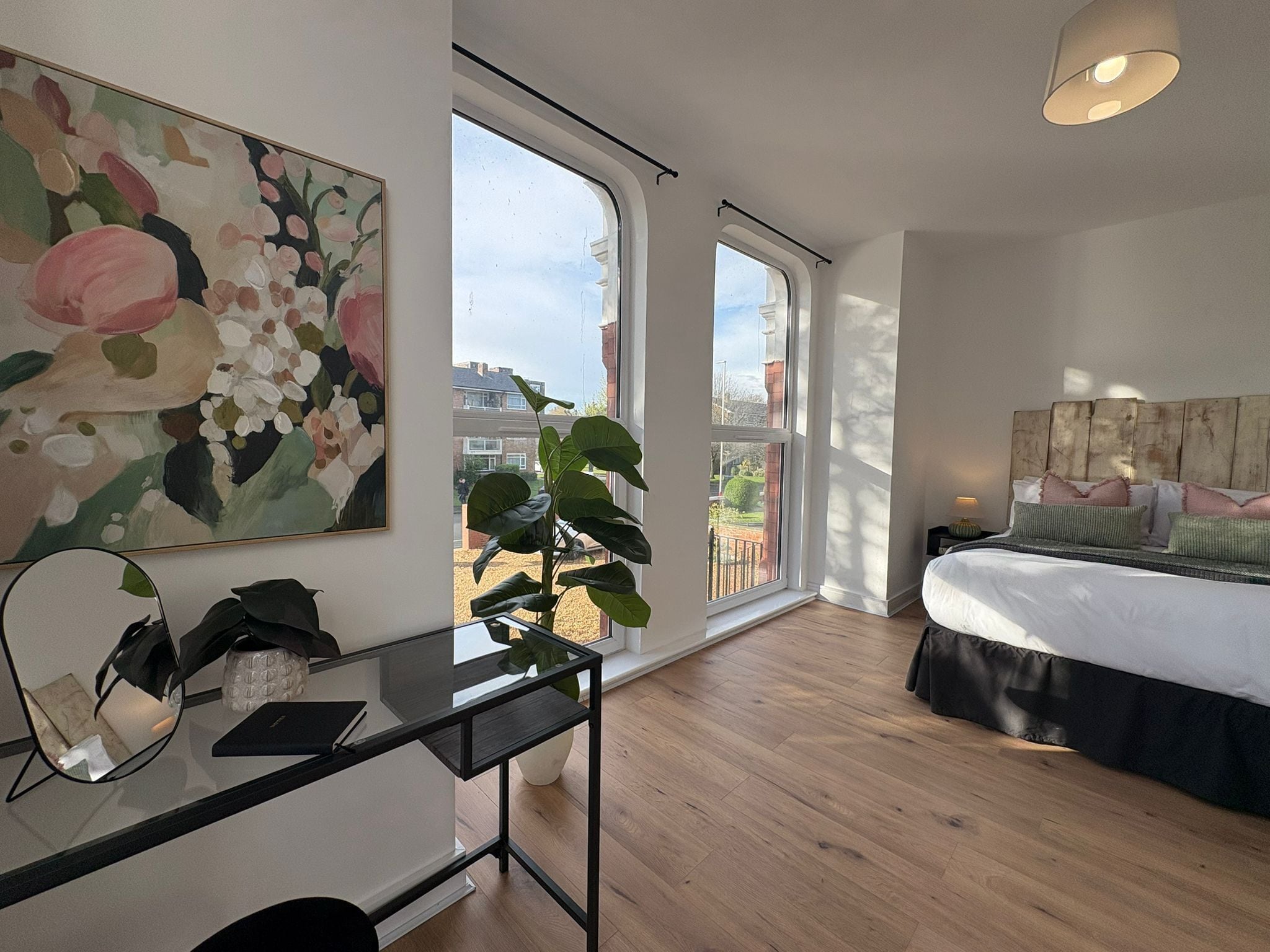 A modern bedroom with a large bed, a desk with a mirror, a potted plant, and a painting on the wall.