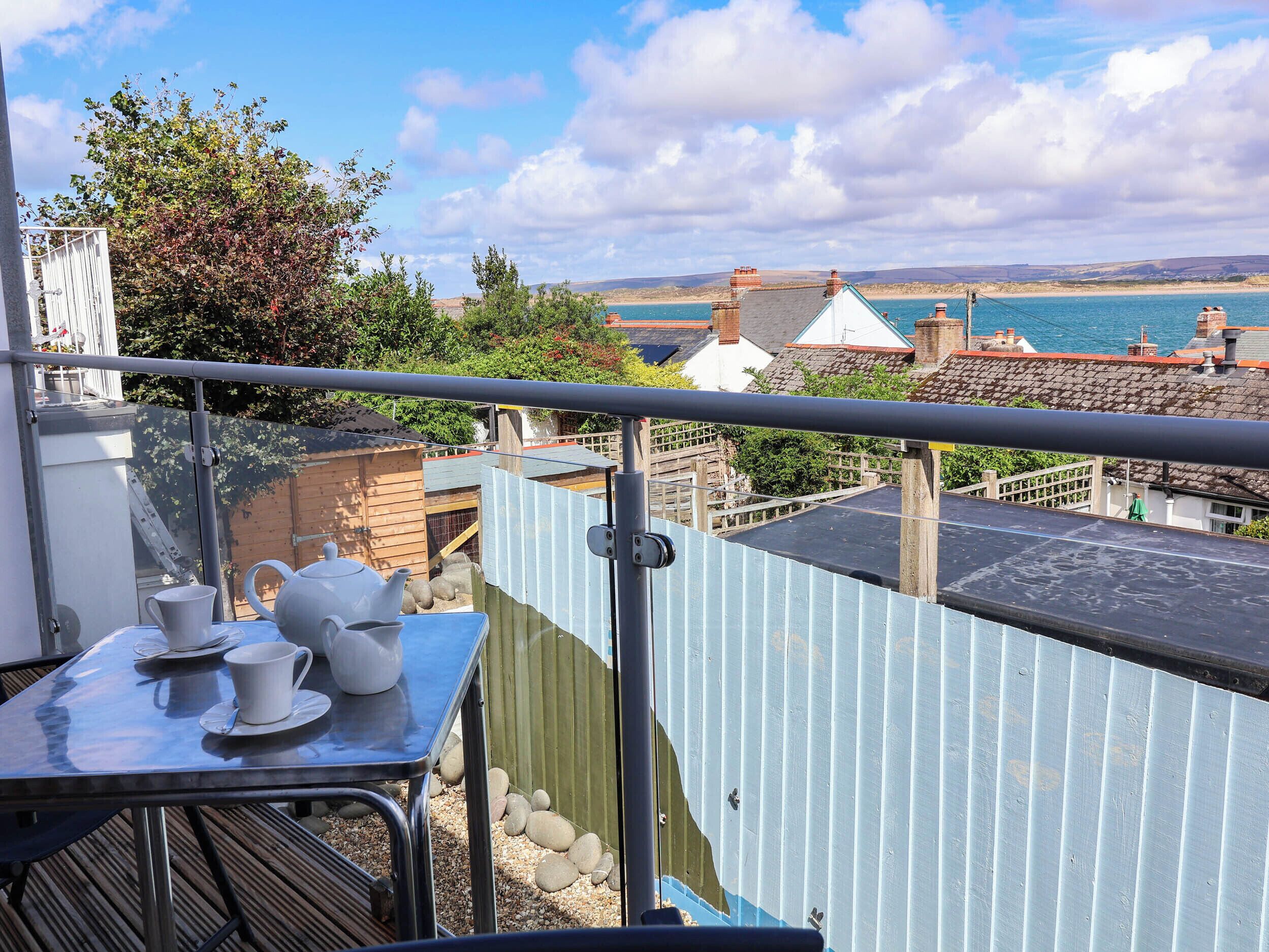 A balcony with a table set for tea, overlooking a coastal landscape with buildings and a body of water.