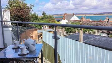 A balcony with a table set for tea, overlooking a coastal landscape with buildings and a body of water.
