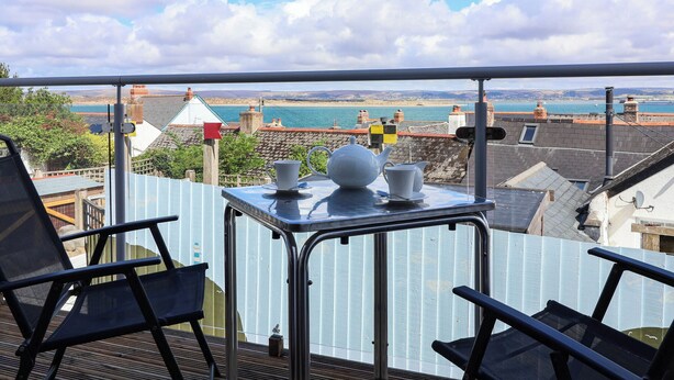 Ein Balkon mit einem für Tee gedecktem Tisch, der Blick auf die Küste mit Gebäuden und ein Gewässer bietet.