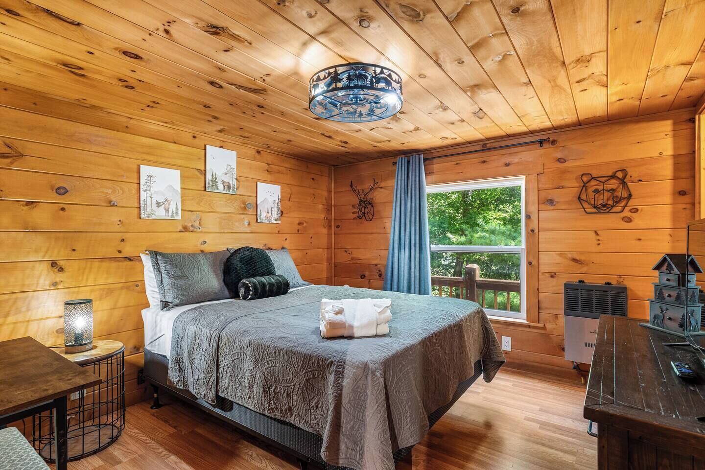 Ein Schlafzimmer einer Holzhütte mit einem Bett, einem Nachttisch, einem Fenster mit Blick auf Bäume und einer Holzbalkendecke.