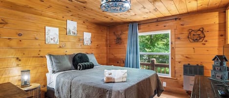A wooden cabin bedroom with a bed, a nightstand, a window with a view of trees, and a wooden ceiling.