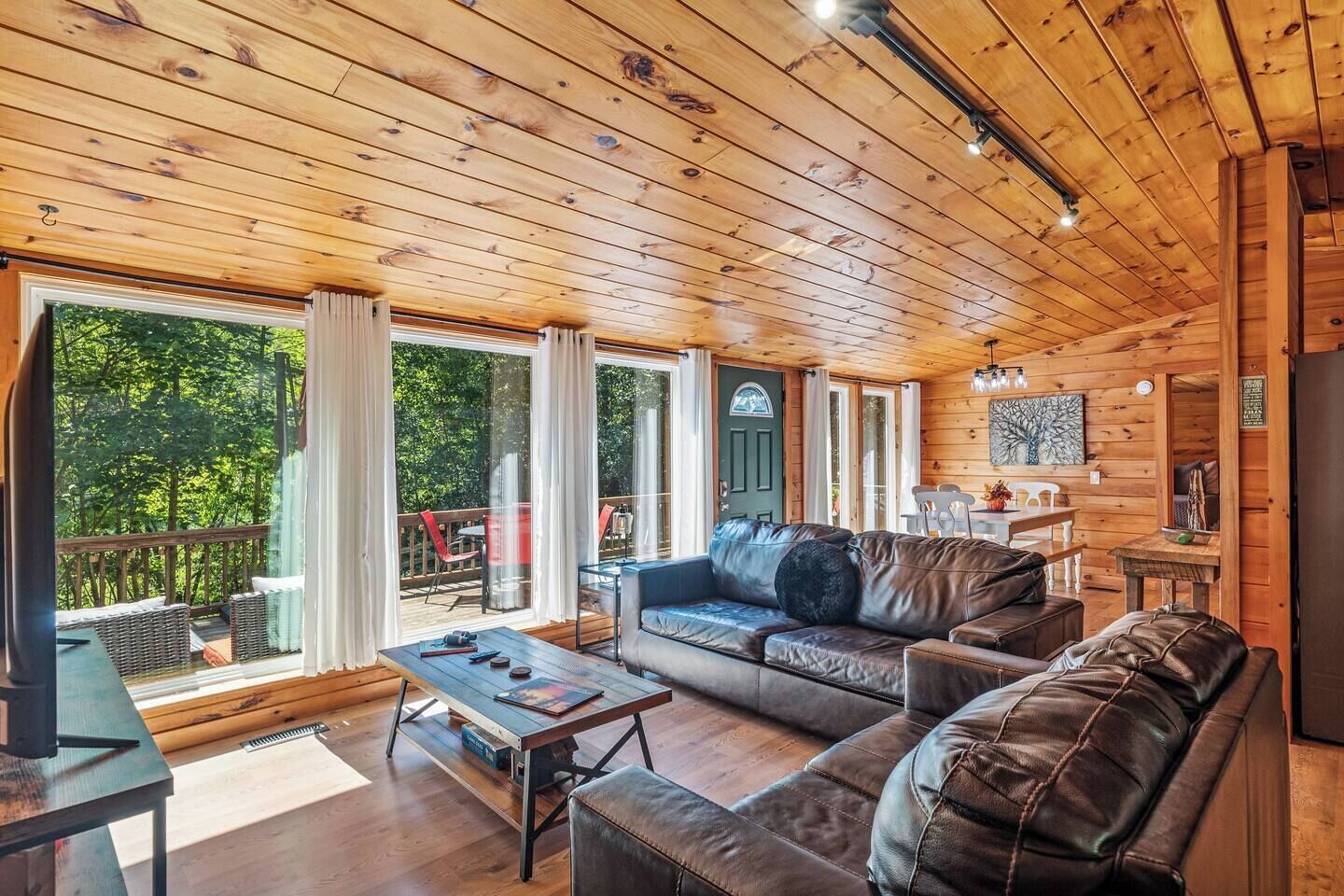 A cabin interior with a wooden ceiling, a sofa, a table, and a view of the outdoors.