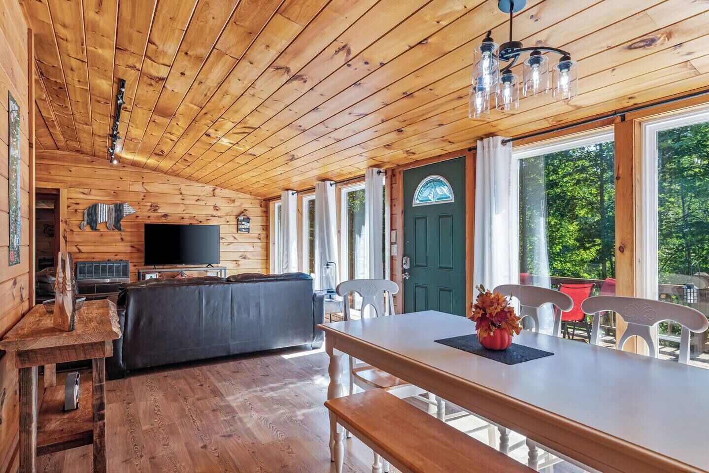 A cabin interior with a wooden ceiling, a television, a sofa, a dining table, and a view of the outdoors.