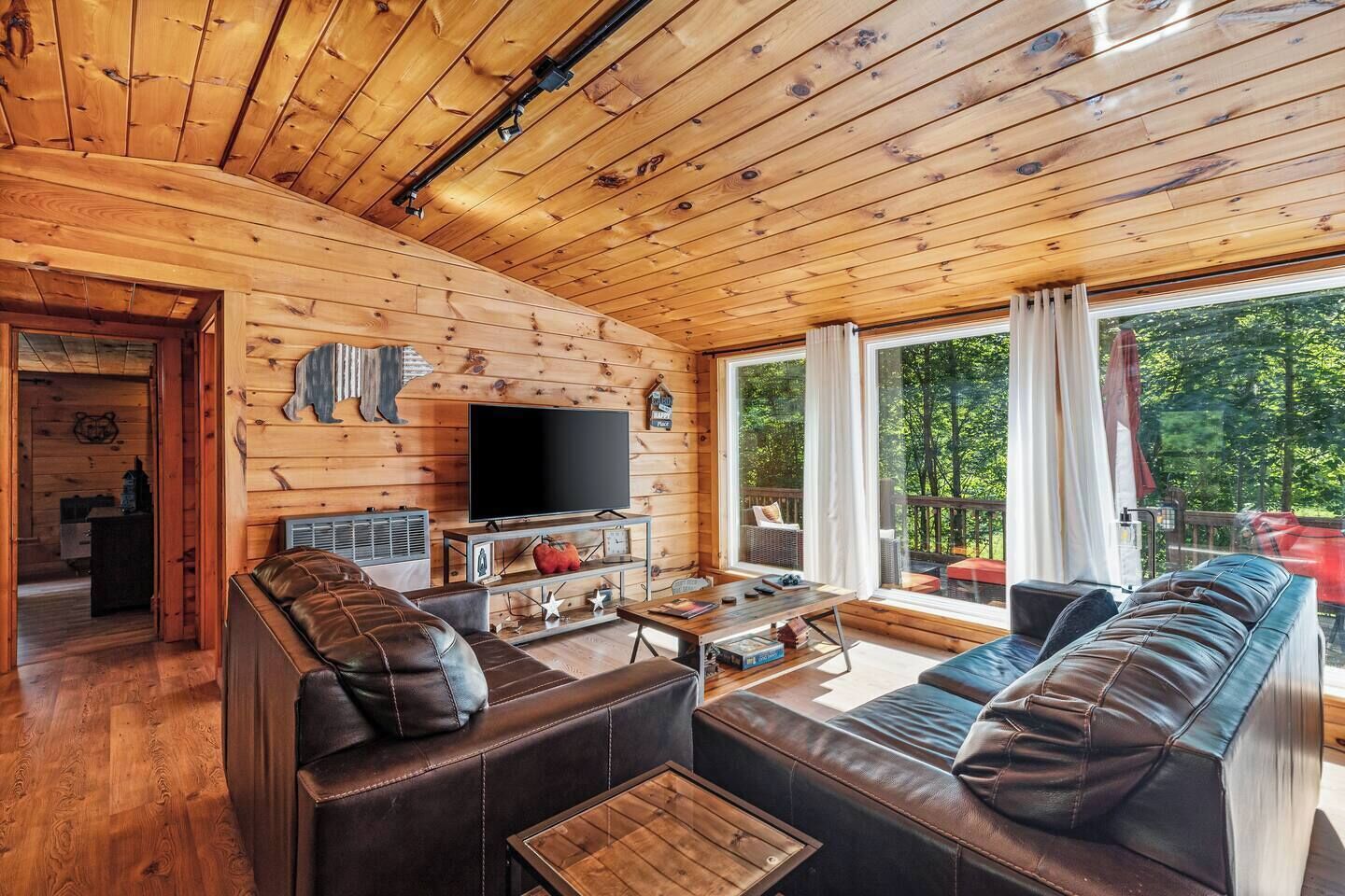 Een gezellig cabin interieur met een houten plafond, een televisie, leren banken en een groot raam met uitzicht op de omgeving.
