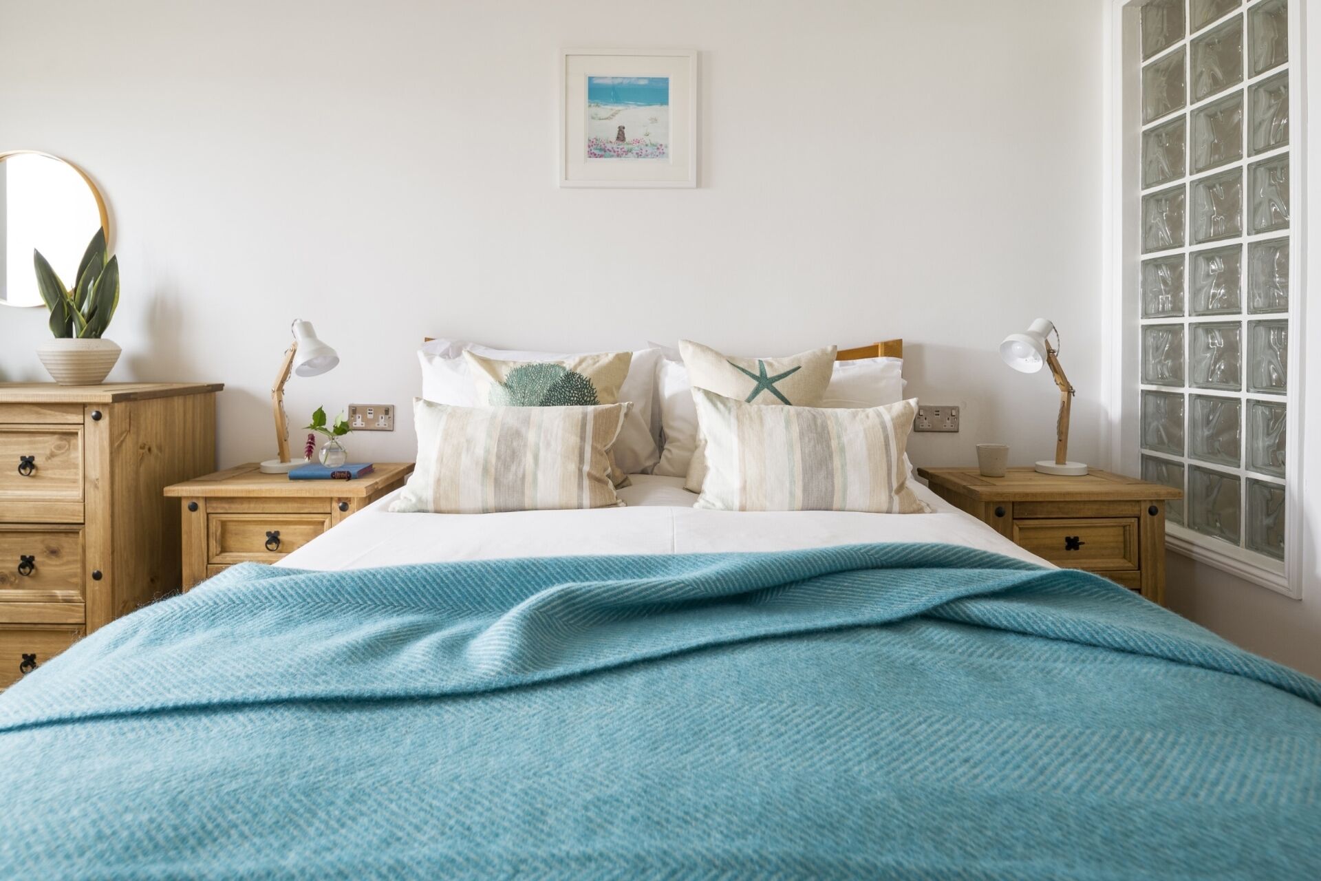 A bedroom with a bed, two bedside tables, a nightstand, a window with frosted glass, a wall-mounted picture, and a potted plant.