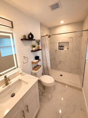 A modern bathroom with a marble floor, a white toilet, a sink with a mirror, and a shower area. - Flora Luxe States (Tampa)
