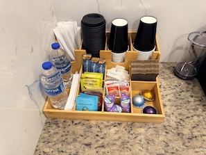 A wooden tray with various items including bottled water, tea bags, coffee pods, and a small decorative object. - Flora Luxe States (Tampa)