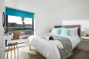 Een moderne slaapkamer met een groot bed, nachtkastje, bureau en uitzicht op een tuin door een schuifdeur.