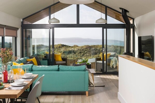 A modern living space with a large glass window, a teal sofa, and a dining area. - Dune Lodge - sleeps 4 guests  in 2 bedrooms (Hayle)