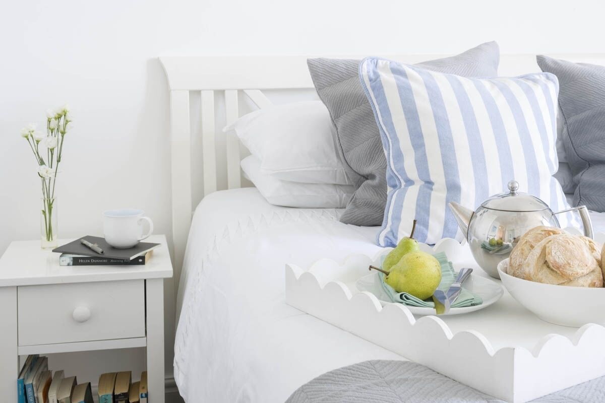 A neatly made bed with pillows, a bedside table with a book, a teapot, a bowl of pastries, and a pear on the bed.
