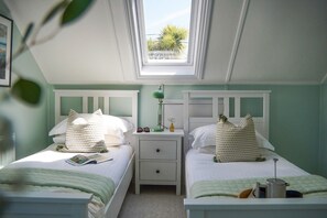 A bedroom with two beds, a nightstand, a lamp, and a window with a view of greenery. - Fourburrows - sleeps 10 guests  in 5 bedrooms (Mawgan Porth)