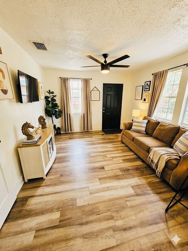 A living room with a wooden floor, a brown sofa, a ceiling fan, and a door. - Beautiful 1-bedroom guest house with AC in Port St. Lucie with Pool (Port St. Lucie)