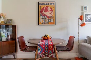 Een eetgedeelte met een ronde tafel, twee stoelen, een kleurrijke tafelkleed, een vintage poster aan de muur en een vloerlamp.